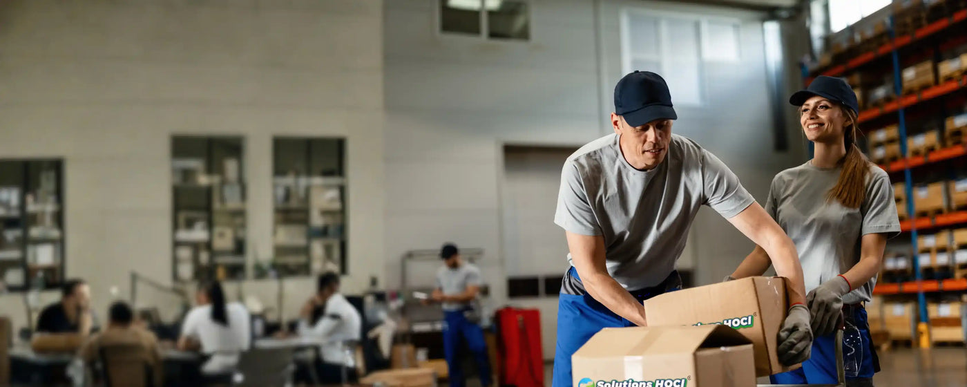 Two workers in a warehouse packaging boxes, with other workers in the background.