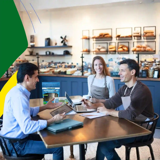 Two men sitting at a table with a woman in a bakery setting, using laptops.