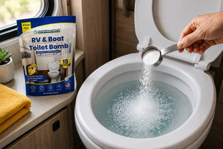A hand pours white powder from a metal scoop into a running toilet bowl, with the package of RV & Boat Toilet Bomb standing nearby on the counter.